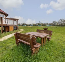 An outdoor wooden table with benches on grass near a wooden house with a porch at Dragonfly Retreat in Upper Marham