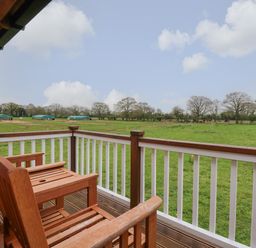 A wooden balcony with two chairs overlooking a grassy field with trees and small structures at Honeybee Retreat in Pentney near Upper Marham