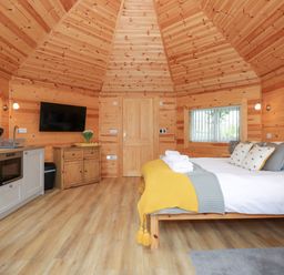 A bedroom with wooden walls and ceiling a bed with pillows and folded towels a kitchenette with a sink microwave and cabinets a wooden cabinet with a vase of flowers a wall-mounted television at Honeybee Retreat in Pentney near Upper Marham