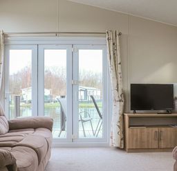 A living room with a sofa a television on a wooden cabinet and glass doors with curtains opening to an outside deck with chairs at Tiptoes Lodge in Carnforth