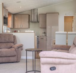 A living area with a brown armchair a beige sofa a small round table and a kitchen with cabinets and appliances at Tiptoes Lodge in Carnforth