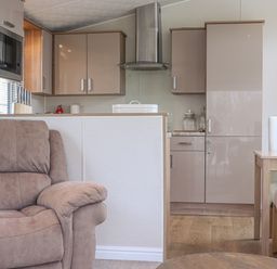 A kitchen with beige cabinets a stainless steel extractor fan a microwave and a knife set next to a living area with a brown armchair at Tiptoes Lodge in Carnforth
