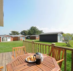 A wooden deck with table and chairs overlooking green grass and small cottages at Little Trebah Atlantic Bays Holiday Park near St Merryn
