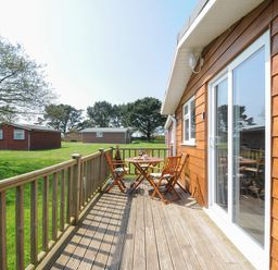 A wooden deck with a table and chairs outside wooden cabins on a grassy area at Little Trebah in Atlantic Bays Holiday Park near St Merryn