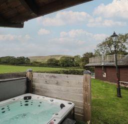 An outdoor hot tub on a wooden deck with a grassy area a wooden cabin a lamppost and hills in the background at Oaks Swanage