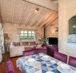 A dining table set with four white bowls and plates and purple chairs in a wooden living room with a beige sofa a television and wood stove at Cherry in Swanage