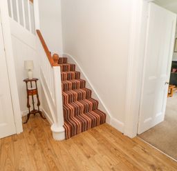 A staircase with striped carpet next to a small wooden table with a lamp and an open door leading to a living room with a fireplace at Crookham Dairy in Crookham near Etal