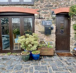 The exterior of a stone building with a dark wooden door and large glass windows with plants in pots and a doormat at Foxglove Barn in South Brent