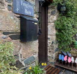 A stone wall with a black mailbox and a door with a knocker next to a shoe rack holding boots at Foxglove Barn South Brent