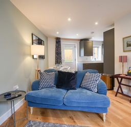 A living room with a blue sofa three cushions a floor lamp a small side table and a small wooden table with a lamp and books at Conybeare Cottage in Lyme Regis