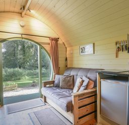 An interior view of a room with a wooden sofa with cushions a small refrigerator a kettle on a countertop knives on a magnetic strip and a round glass door showing an outdoor garden at Pendragon Pod in Axminster