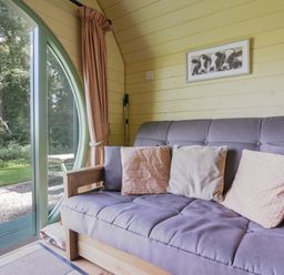A room with a gray sofa and pillows next to a round glass door opening to a garden at Pendragon Pod in Axminster