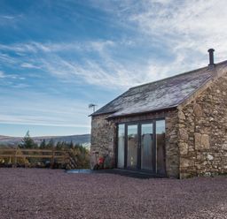 A stone house with glass doors and a chimney on a gravel yard with hills and trees in the background at The Hidden Place in Watermillock near Pooley Bridge