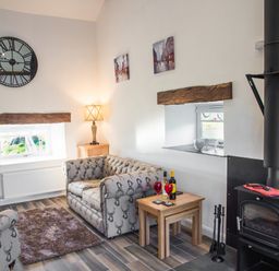A living room with two armchairs a wood burning stove a side table with wine glasses and bottles a lamp a wall clock and two small windows at The Hidden Place in Watermillock near Pooley Bridge
