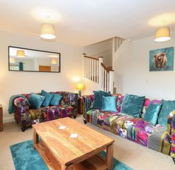 A living room with patchwork sofas, teal cushions, wooden coffee table, side tables with lamps, and a staircase at 30 Burtons Mill in Stalham