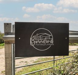 A metal gate with a black sign showing a house and the text Binneford Holiday Lodges in a rural outdoor setting