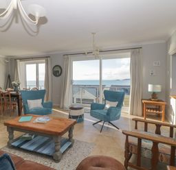 A living room with blue chairs a wooden coffee table and a dining area with a sea view at Looe Island View in Downderry
