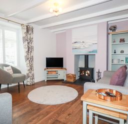 A living room with a gray armchair, a sofa with purple pillows, a round rug, a TV on a stand, a fireplace with a stove, and a shelving unit at Still Waters in Pentewan