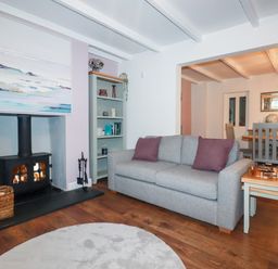 A living room with a grey sofa and purple pillows next to a fireplace and a bookshelf with a dining area visible in the background at Still Waters in Pentewan