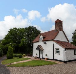 A white cottage with a red tiled roof and a chimney surrounded by grass and trees at Grove Cottage in Leominster