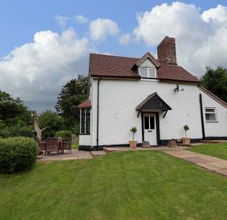 A white cottage with a red roof and chimney on a grassy lawn with a paved pathway and outdoor seating at Grove Cottage in Leominster