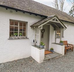 An entrance door with a small porch and potted plants next to windows and a wooden bench outside a white textured cottage