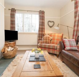 A living room with a wood burning stove basket with logs checkered armchair brown leather sofa wooden coffee table and windows with checkered curtains at Larch Cottage at Esthwaite Water in Hawkshead