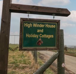 A wooden sign for High Winder House and Holiday Cottages on a post beside a rural road with fields and a wire fence