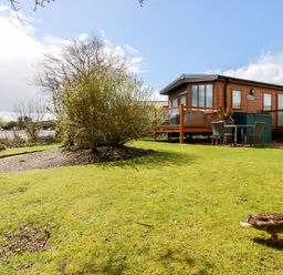 A wooden cabin with a deck and outdoor table and chairs on a grassy lawn with bushes and a duck at Lake View 10 in Par
