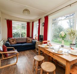 A living room with a blue sofa red curtains a wooden dining table and large windows overlooking greenery at Eweleaze in West Bexington