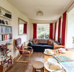 A living room with a blue sofa red curtains a wooden chair table set with food and books on a shelf at Eweleaze in West Bexington
