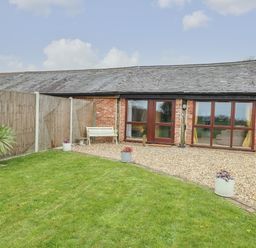A backyard with grass, potted plants, a bench, gravel area, and a brick building with wooden doors and windows at Bee Happy Barn in Beighton near Acle