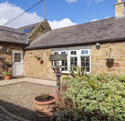 A courtyard with gravel and potted plants in front of a stone house with white windows and a door at West Farm Cottage in Longhorsley