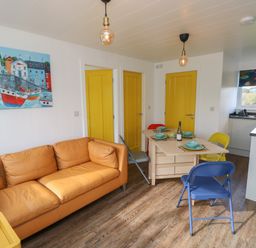A living room with a tan sofa yellow doors a wooden dining table with colorful chairs and a kitchen area at Nanny's Little House in Sandown