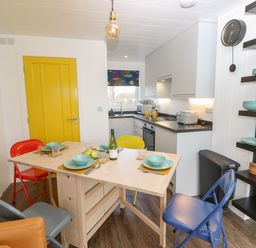 A kitchen with a wooden table set for four, colorful chairs, yellow doors, appliances, and wall shelves with plates at Nanny's Little House in Sandown