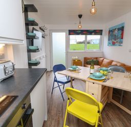 A kitchen and dining area with a toaster and induction stove a wooden table set with plates bowls and glasses yellow and blue chairs and a brown sofa near a window at Nanny's Little House in Sandown