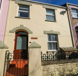 A cream colored terraced house with two windows and a wooden door behind a black wrought iron gate at Stones Throw in Tenby
