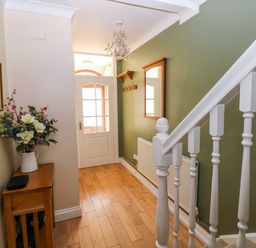 A hallway with wooden floor a staircase with white balusters a wooden table with a flower vase a green wall with a mirror and coat hooks at Stones Throw in Tenby