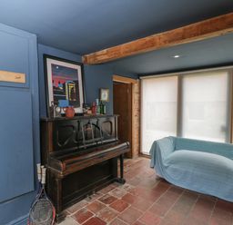 A room with a black upright piano decorated with bottles and a framed picture and a blue covered sofa near large windows at Brandeers Long Barn in Brandier near Minety