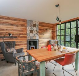 A dining room with a wooden table, mixed chairs, wood-paneled walls, a fireplace, and large windows showing greenery at The Big Box Brandeers in Brandier near Minety