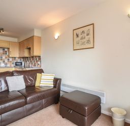 A living room with a brown leather couch, striped cushions, a brown ottoman, a small kitchen with tiled backsplash, and a framed picture on the wall at 3 Bay View Court in Lyme Regis