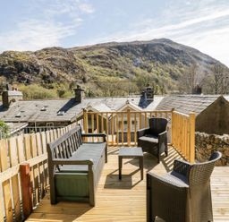 An outdoor wooden deck with chairs a bench and a small table overlooking rooftops and a hillside at Henfro in Beddgelert