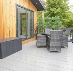 An outdoor deck with grey wicker chairs around a table and a black storage box at Watch Tree Lodge in Allithwaite near Cartmel