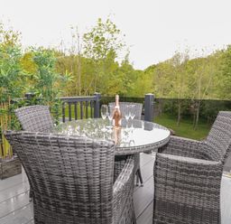 A balcony with a round glass table surrounded by four woven chairs and a bottle with glasses near plants at Watch Tree Lodge in Allithwaite near Cartmel
