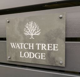 A sign with a tree illustration and text watch tree lodge on a wooden surface at Watch Tree Lodge in Allithwaite near Cartmel