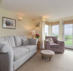 A living room with a grey sofa armchair round footstool side table lamp and flowers at Chase View Bowland Bridge near Bowness-On-Windermere