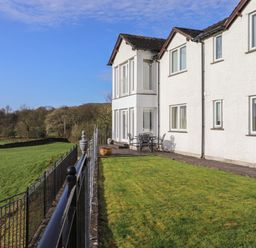 A white house with multiple windows and a metal patio table with chairs on a paved patio beside a green lawn and black metal railing at Chase View in Bowland Bridge near Bowness-On-Windermere