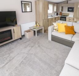 A living room with a large gray sofa with yellow and orange cushions a TV on a stand with a fireplace below a small table with candles and magazines and a kitchen area with wooden cabinets at The Waves Little Haven near Broad Haven