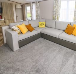 A living area with a sectional sofa with colorful cushions near windows with patterned curtains and a kitchen with wooden cabinets at The Waves in Little Haven near Broad Haven