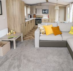 A living room with a grey carpet sectional sofa with yellow pillows a beige side table and a kitchen with wooden cabinets at The Waves in Little Haven near Broad Haven